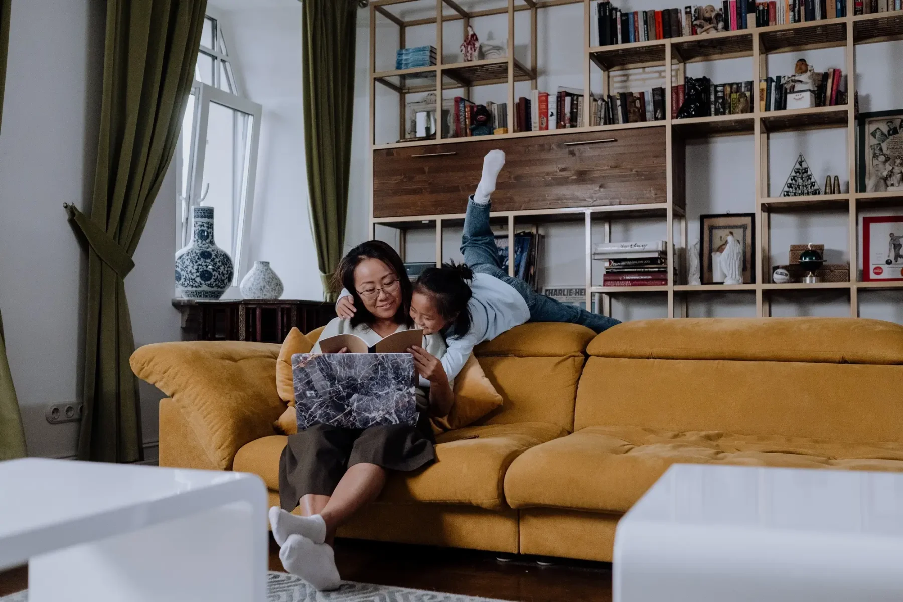 A young girl climbing over the back of a couch to embrace her mother who is reading a book and working on a laptop