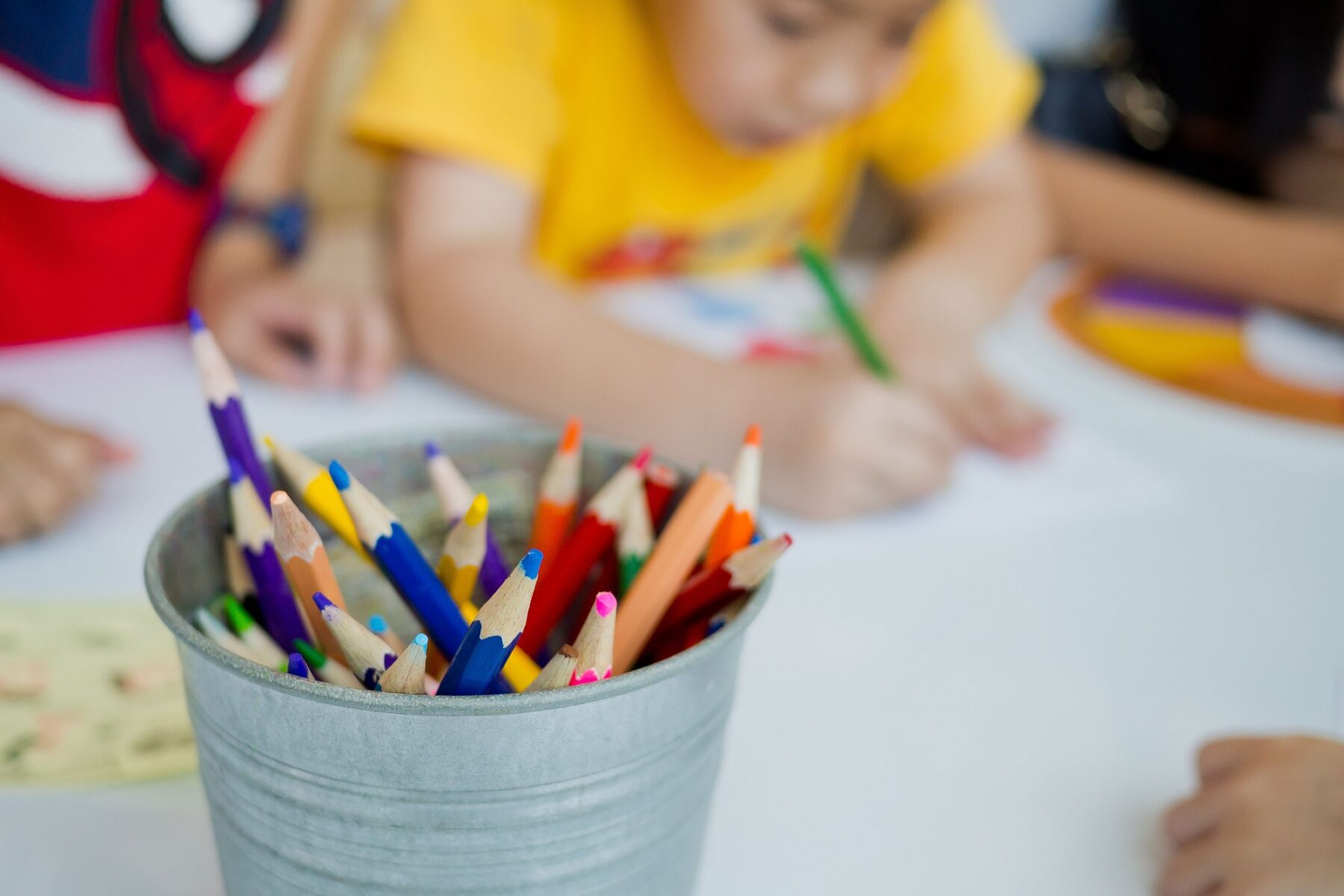 Colored pencils in a small bucket. Children coloring at a table.